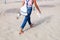 A girl in jeans walks on the sand barefoot, holding sandals in her hands, rear view