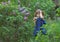 A girl with long hair 3 years old in the spring stands next to lilac bushes in the green grass in a blue dress with a camera in he