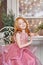 Girl with long red hair in a pink dress sitting at the table. Carnival holiday birthday. Portrait of a cheerful red-haired girl
