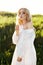 Girl in a long white dress with a dandelion flower in her hand stands in grass in a field. Blonde woman in the sun in a light