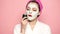 A girl with a mask on her face and a towel on her head eating a cucumber on a pink background. Beautiful girl with