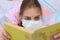 Girl in a medical mask reads a book while lying in bed, close-up