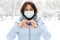 A girl in a medical mask in the winter outdoors on a background of snow makes a heart with her hands.