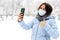 A girl in a medical mask in the winter outdoors on the background of snow makes a selfie on the phone.