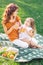 Girl and mommy drinking orange juice from plastic cups sitting on the blanket in the park together