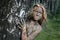 A girl with a mud mask peeps out from behind a birch trunk at an ecological resort