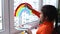 Girl painting rainbow on window during quarantine at home.