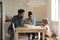 Girl Paints At Kitchen Table As Parents Look At Laptop