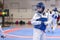 A girl participating in Taekwondo competitions in a blue protective vest and a protective helmet is a duel in Taekwondo against th