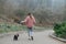 Girl in a pink coat walks the alley with a small dog on a leash, the view from the back. Woman walks with dog in spring gray park