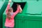 A girl in a pink jacket, throws a paper bag, in a green garbage container, there is a place for the inscription, rear view