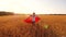 Girl plays Superhero, child runs in red cloak across field with wheat under the blue sky. Slow motion.