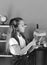 Girl with ponytails stands by pile of books and bookshelf. Schoolgirl with proud face holds pink piece of paper