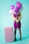 Girl in purple dress with feathers holding balloons in front of face near huge gift box on turquoise