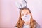 Girl in rabbit bunny ears on head and protective mask with colored eggs on pink background. Covid Easter holiday