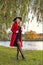 A girl in a red coat and hat stands under the branches of a willow on the bank of the river of the lake