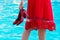 Girl in a red dress and with red shoes in hand at the pool_