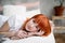 The girl with red hair is on  bed and looking forward, her hands are folded under her head, a white bed, is at home, self-