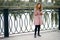 A girl with red hair in a pink checkered dress stands on the embankment and looks at her phone in a funny video