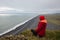 A girl in a red jacket sits on a cliff above the sea shore with black lava sand stretching to the horizon