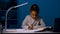 The girl rewrites the task from the textbook into a notebook. A schoolgirl sits at a table by the light of a night lamp