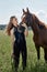 Girl rider stands next to the horse in the field. Fashion portrait of a woman and the mares are horses in the village in the grass