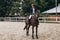 A girl rides fast on a brown racehorse next to a hurdle at a show jumping competition on a clear summer day