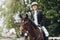 A girl rides fast on a brown racehorse next to a hurdle at a show jumping competition on a clear summer day