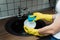 A girl in rubber gloves foams soap with a sponge on a dirty plate against the background of an empty sink. Housekeeper is going