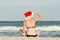 Girl in Santa hat with the inscription New Year on the back is sitting on the beach and looks into the distance