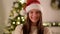 Girl in Santa Hat with Long Dark Hair Posing on Christmas Tree Background. Happy and Emotional Woman Smiling at the