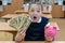 Girl schoolgirl holds a piggy bank in her hands and fake dollars in a school classroom. The concept of expensive education and