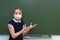 A girl a schoolgirl in a medical mask stands at the blackboard and shows her hands on free space. The emotion of surprise and