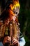 Girl with shamanic feather mask and historic dress in woodland surroundings playing wooden ornamental flute.