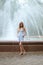 Girl in short blue dress stands in front of fountain in summer park