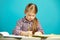 Girl sits at desk and writes to notebook, on blue isolated background. First grader in school.