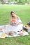 Girl sitting on plaid near fruits and hat, eating watermelon, grass in background.