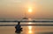 Girl sitting by the sea at sunset and looking a passing boat, in light summer dress. She looks the Sun sets over the horizon.