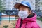 A girl sitting on a seesaw on playground near high-rise buildings with apartments, a medical mask on her face protects against