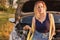 Girl is sitting with a tow rope on the hood of a broken car on a rural road in the rays of the sunset