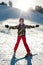 A girl skier in red pants and a green jacket on skis stretching out her arms happy with the sun`s rays