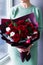 A girl standing near the window in a sea-colored dress holds in her hands a burgundy bouquet with red roses with white flowers and