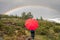 A girl standing under a beautiful rainbow and dark skies with a red umbrella in the middle of a green natural landscape scenery wi