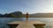 Girl standing up with a stalk hat on and red dress with her arms up looking at the mountains with her shoes on a dock by a lake