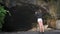 Girl stands in front of entrance to a large cave from which bats fly out and takes video on her phone. A woman in denim