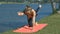 Girl Stands in Hard Yoga Position on Lake Bank Grass