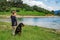 Girl stands near dog of Rottweiler breed in meadow next to lake, against hilly valley with spruce forests