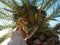 A girl stands under a spreading palm tree looking up and holding on to a thick trunk. Photo from bottom to top