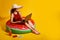 girl in straw hat sits in inflatable watermelon ring uses pc on yellow background. travel concept