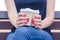 Girl on the street sits on a bench during the day. in hands with red manicure holds 2 disposable cups with tasty aromatic coffee.